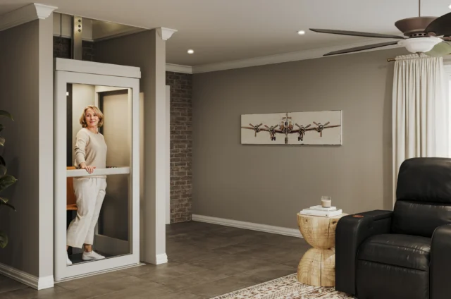 Woman in white clothing stands in her residential elevator parked in her living room with a black leather chair.