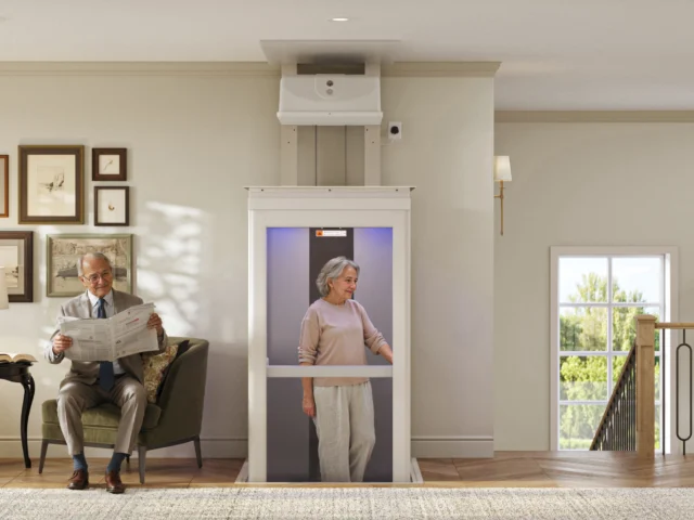 Older woman riding in her elevator with her husband sitting on the couch with a newspaper