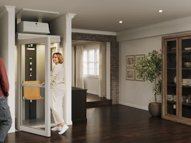 Woman in white clothing enters her Pollock elevator, parked in a living room with dark wood floors, some paintings on the wall, and a small fern beside the paintings.