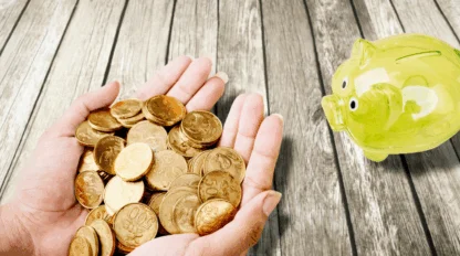 Cupped hands holding gold coins over a wooden table, offering them to a yellow, plastic piggy bank