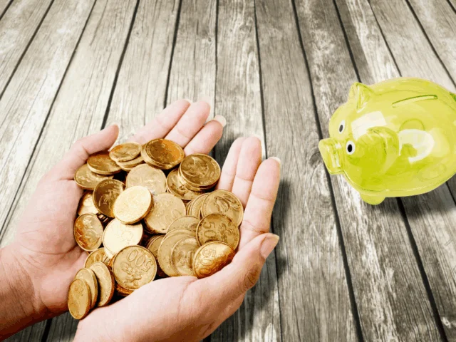 Cupped hands holding gold coins over a wooden table, offering them to a yellow, plastic piggy bank