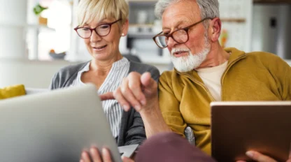 Older couple with white hair and dark glasses are reading off a laptop and tablet screen in a well lit home.