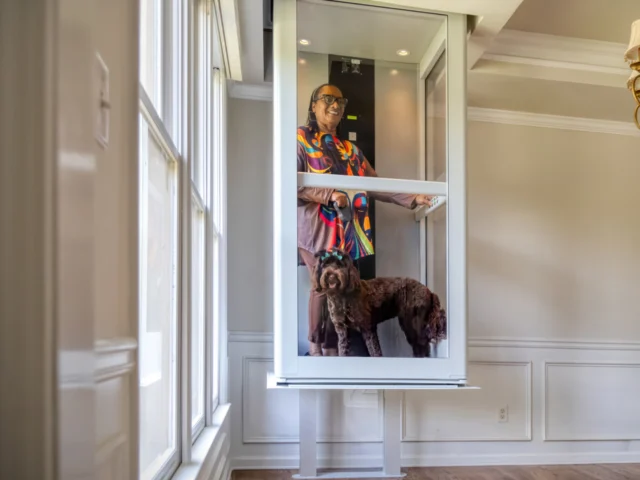 Homeowner riding a compact glass residential elevator with her dog inside a bright home interior, illustrating accessible aging-in-place mobility.