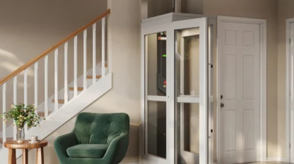 Compact shaftless home elevator installed beside a staircase in a modern living room, featuring glass panels, white framing, and a small footprint design for residential accessibility.