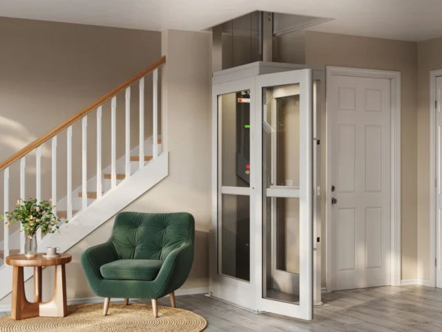 Compact shaftless home elevator installed beside a staircase in a modern living room, featuring glass panels, white framing, and a small footprint design for residential accessibility.