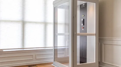 Modern white shaftless residential elevator with glass panels installed in a bright home interior with hardwood floors and large windows.