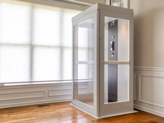 Modern white shaftless residential elevator with glass panels installed in a bright home interior with hardwood floors and large windows.