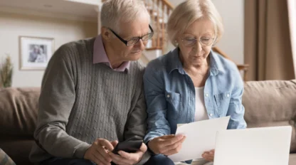 An older couple sitting on a couch with stairs behind them while reading a piece of paper with their laptop and phone.