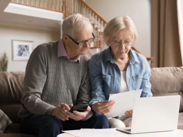 An older couple sitting on a couch with stairs behind them while reading a piece of paper with their laptop and phone.