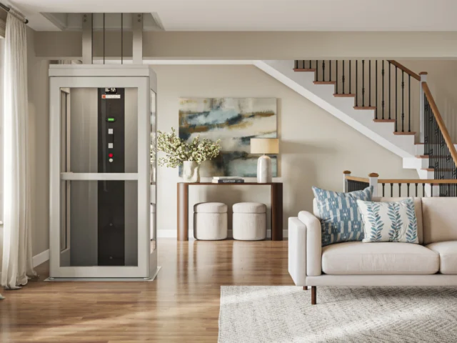 A white home elevator parked in a bright, decorated living room with stairs leading to the second floor in the background.