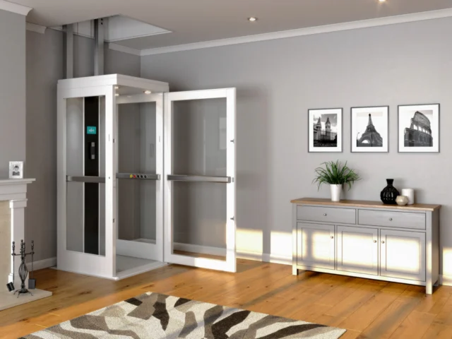 A while home elevator sits in the corner of a well lit room with grey walls and a hardwood floor, its door open.